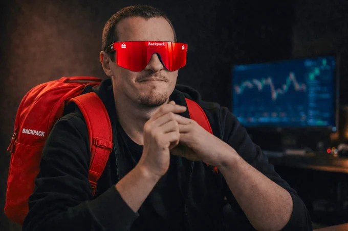 A person wearing red-tinted sunglasses and a red backpack sits confidently, with a digital display of charts in the background.