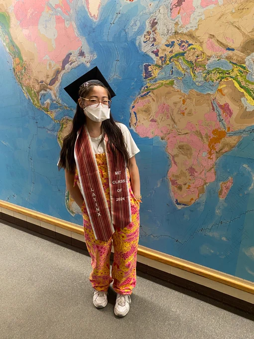 Author of tweet standing in front of a world map on a wall. She wears a black graduation cap, pink and yellow overalls with a whimsical bright print, a red graduation stole that says LATINX and MIT Class of 2024. Her N95 duckbill respirator is white, and she wears amber framed glasses.