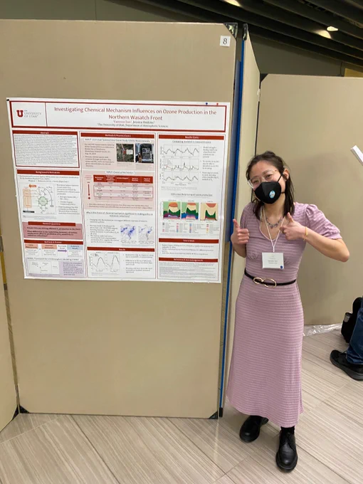 Author of post, an Asian American woman, standing next to her conference poster giving two thumbs up. She wears a long ankle-length purple dress, a black belt with gold buckle at the waist, black Oxford style shoes, and gold rimmed cat eye glasses. Her N95 respirator is black and her brown hair is half tied up. 