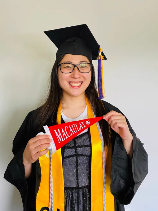 Author of tweet standing and holding a small red colored pennant that says Macaulay and in small letters, a white logo that says CUNY. She wears a graduation robe, cap, and a black sheer dress with golden sparkles, and a bright golden yellow graduation stole