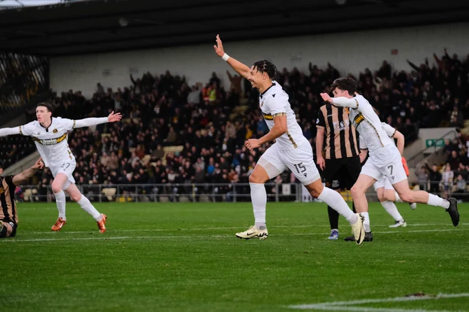 A group of soccer players in white uniforms celebrate scoring a goal on the field. The mood is triumphant and energetic, with a crowd cheering in the background.