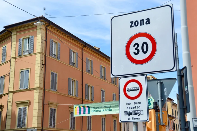 People of all ages and abilities walk, cycle, drive, and ride public transportation along the low-traffic streets of Bologna, which are lined with buildings, trees, bike infrastructure, and prominent 30 km/h speed limit signage and road markings.