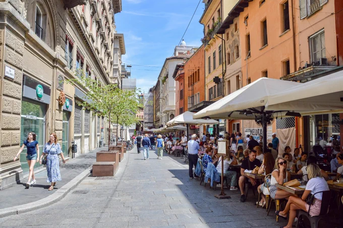 People of all ages and abilities walk, cycle, drive, and ride public transportation along the low-traffic streets of Bologna, which are lined with buildings, trees, bike infrastructure, and prominent 30 km/h speed limit signage and road markings.