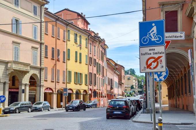 People of all ages and abilities walk, cycle, drive, and ride public transportation along the low-traffic streets of Bologna, which are lined with buildings, trees, bike infrastructure, and prominent 30 km/h speed limit signage and road markings.