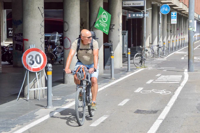 People of all ages and abilities walk, cycle, drive, and ride public transportation along the low-traffic streets of Bologna, which are lined with buildings, trees, bike infrastructure, and prominent 30 km/h speed limit signage and road markings.