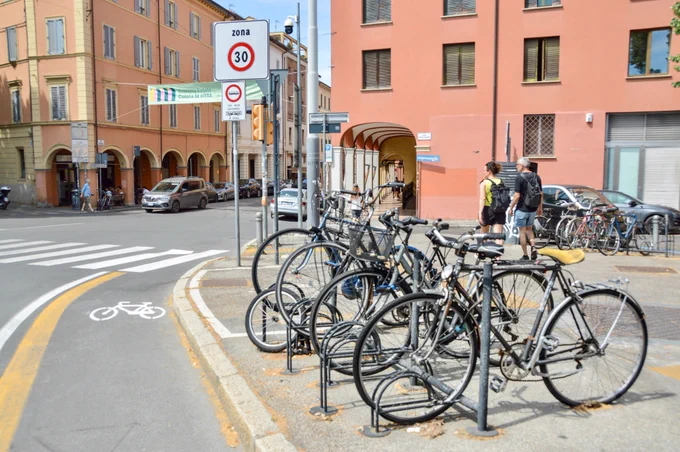 People of all ages and abilities walk, cycle, drive, and ride public transportation along the low-traffic streets of Bologna, which are lined with buildings, trees, bike infrastructure, and prominent 30 km/h speed limit signage and road markings.