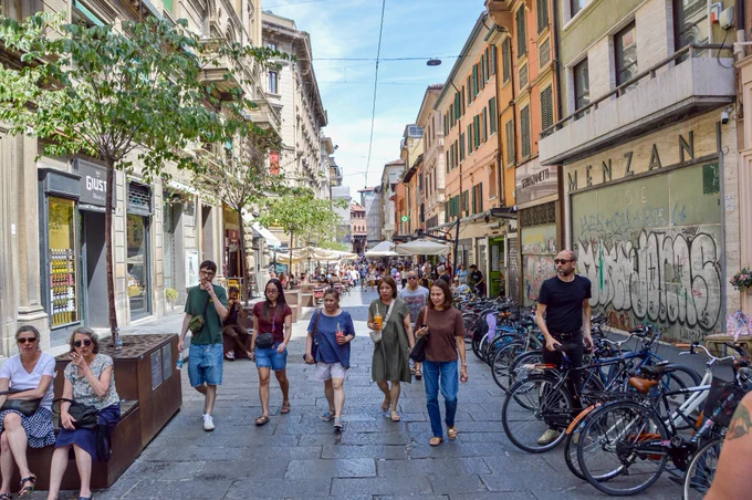 People of all ages and abilities walk, cycle, drive, and ride public transportation along the low-traffic streets of Bologna, which are lined with buildings, trees, bike infrastructure, and prominent 30 km/h speed limit signage and road markings.