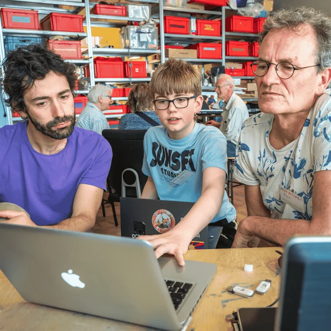Un mozo con camiseta azul apunta á pantalla dun laptop aberto que está sobre unha mesa, acompañado de dúas persoas. Na parede detrás deles atópanse estanterías organizadas ordenadamente.