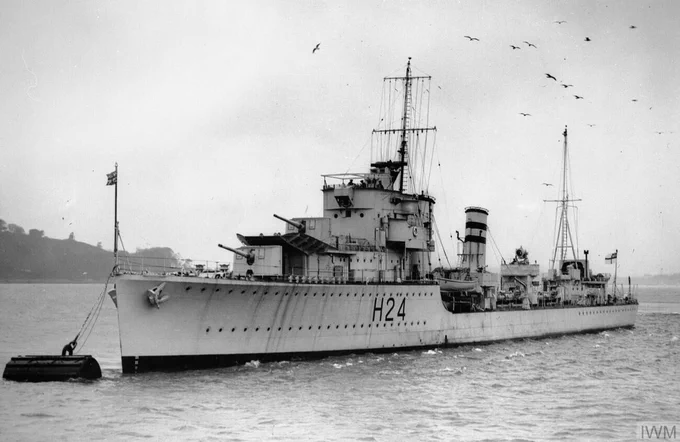 Royal Navy destroyer HMS Hasty. Image: Imperial War Museum FL 13795. Black and white photograph of a small warship moored to a buoy in harbour, with the shoreline visible behind and a lot of seagulls overhead.