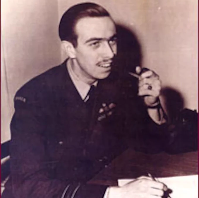 Byron Rawson sits at a desk in a dark uniform jacket, holding a telephone receiver in one hand and a pen over paper with the other. He has neatly combed dark hair and a thin moustache. The sepia-toned photo is framed closely, with a plain wall behind him and a shadow cast to one side.

