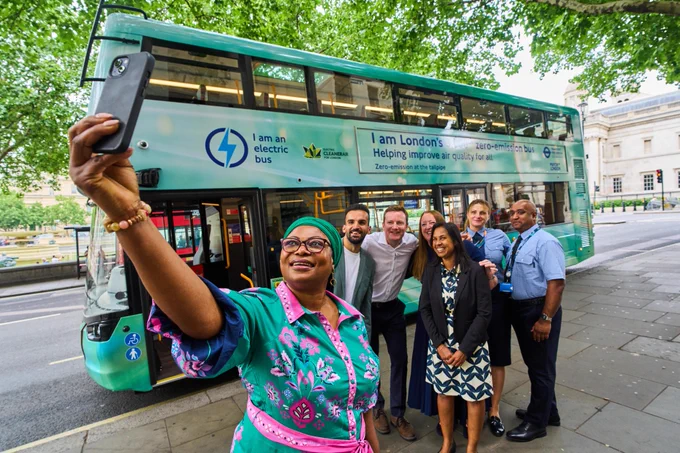 \"Bus Aunty\" in a green outfit takes a selfie with six smiling adults from TfL and Stagecoach, including bus staff, in front of a green double-decker zero emissions bus. Text on bus: 'I am an electric bus. I am London's zero-emission bus. Helping improve air quality for all. Zero-emission at the tailpipe.'