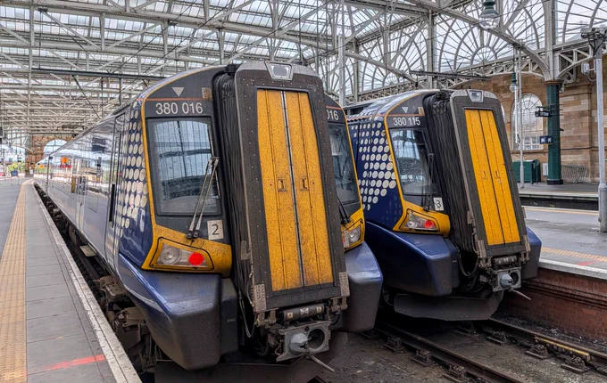 Two dark blue ScotRail trains are parked side-by-side at a railway station platform under a large, arched glass and steel roof structure. Both trains feature prominent, textured yellow gangway doors on their fronts and yellow trim around the driver's cabs. The train on the left is numbered 380 016 on its front window, while the train on the right is numbered 380 115. The side of the trains displays the white dotted Saltire graphic livery. The platform to the left shows a textured yellow tactile paving strip.