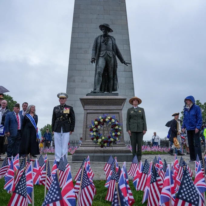 A member of the military in black and white suite and park ranger in green suite stand at attention in front of the Colonel William Prescott statue and the Bunker Hill Monument. A wreath is on a stand in between them and there are American and British flags in a flag garden in the foreground. 