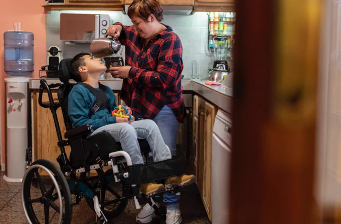 A caregiver stands in a kitchen helping a child seated in a wheelchair. The child looks up at the caregiver while receiving assistance. Kitchen cabinets, countertops, and household items are visible in the background, emphasizing a daily family caregiving setting.
