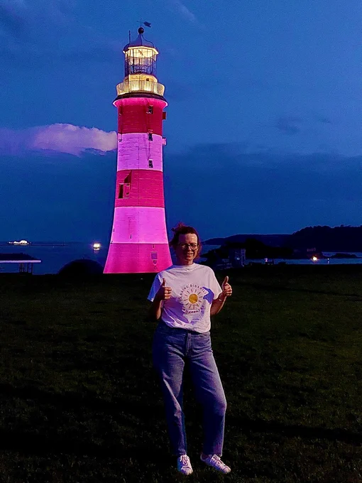 Woman with thumbs up stood in front of a red and white striped lighthouse which is lit up in purple