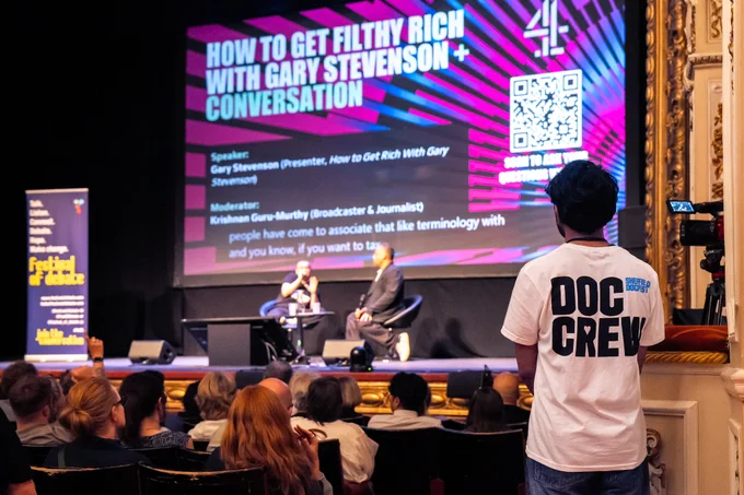 A photo of two people sat onstage in conversation, in an old-fashioned theatre. In the foreground is a person watching them, wearing a white t-shirt with "DOC CREW" printed on the back