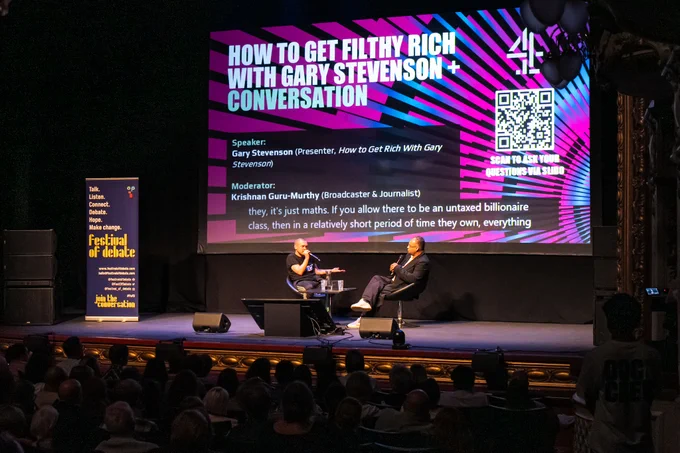 A photo of two people sat onstage in conversation, in front of a big screen with the words "HOW TO GET FILTHY RICH WITH GARY STEVENSON   CONVERSATION"