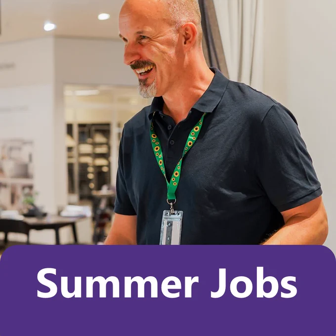 A person wearing a dark polo shirt and a sunflower lanyard badge stands in a bright indoor workplace, smiling while looking to the side. The background shows a clean, modern work environment with tables and equipment slightly out of focus. 

Large text at the bottom of the image reads “Summer Jobs”.

Image Credit: Business Disability Forum