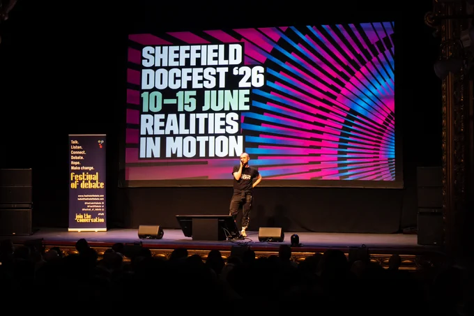 A photo of a man stood onstage with a microphone, in front of a big screen with the words "SHEFFIELD DOCFEST '26, 10–15 JUNE, REALITIES IN MOTION
