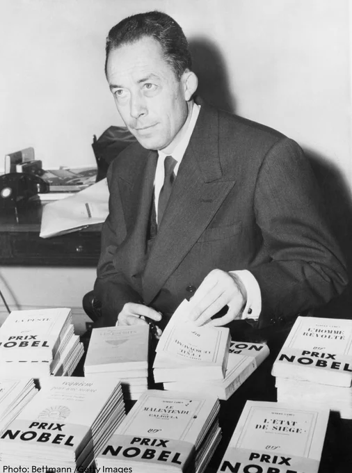 Albert Camus sits at a desk, wearing a suit surrounded by stacks of books labeled "Prix Nobel"