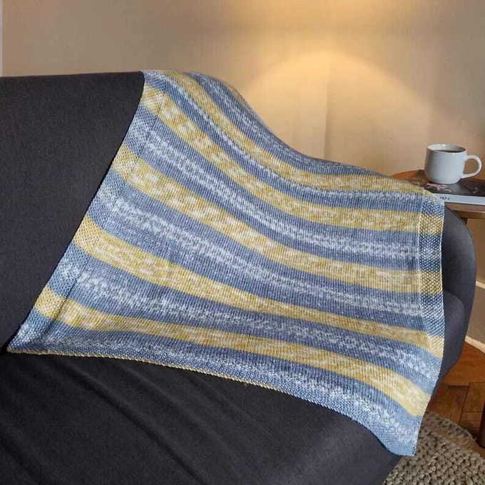 A mustard, grey and cream striped hand knitted blanket is draped over a dark grey armchair. A lit lamp glows against a neutral wall. A small wooden side table holds a white mug and a book. Wooden flooring and a textured rug are visible below.