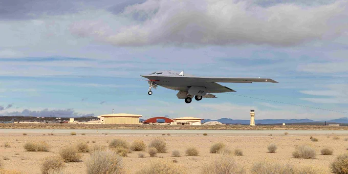 A B-21 Raider flying low over a desert airfield with sparse bushes and a control tower in the background.