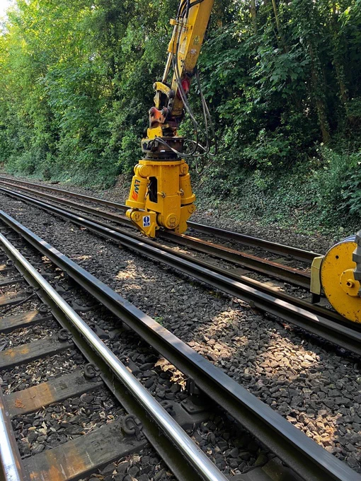 A Road, Rail Vehicle (RRV) gripping and aligning steel rails on a track, surrounded by dense green vegetation in a cutting.