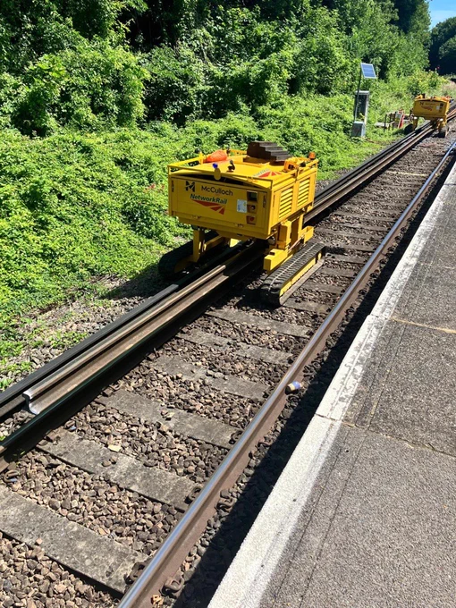 Yellow rail maintenance machine on railway tracks beside a station platform, with green vegetation lining the track.