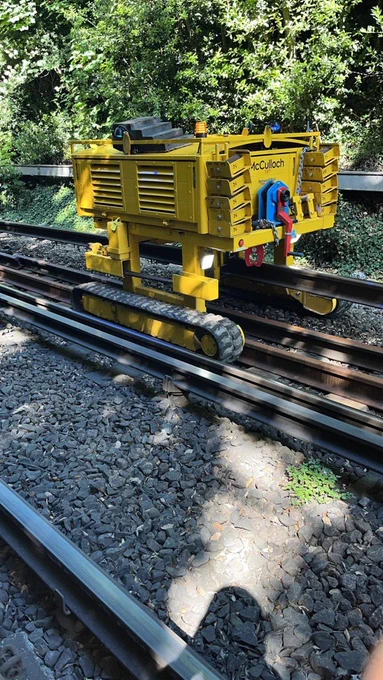 Close-up of a yellow rail-mounted maintenance machine positioned on tracks in a leafy cutting, showing mechanical components and rails in detail.