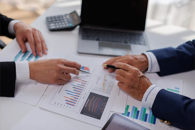 Two people are discussing various business charts and graphs on a table, which include bar and line graphs. A laptop, a calculator, and a pen are also visible.