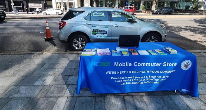 Outdoor Mobile Commuter Store table with transit brochures and a laptop beside a parked silver car on a city sidewalk.