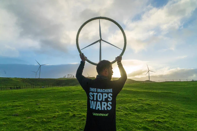 Someone holds a circle up to frame a wind turbine in the distance, making it look like a giant peace sign. They're wearing a t-shirt with the message: "This machine stops wars"