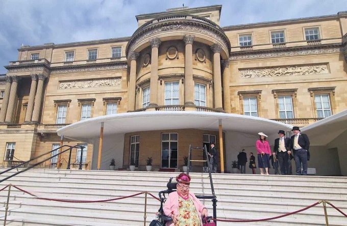The image shows Penny sitting at the bottom of white steps which are cordoned off with red rope, at the back of Buckingham Palace, which is a brown and dull stately home building with tall windows and a centre half-dome with pillars. At the top of the steps are three figures in formal wear. It is a sunny day with light clouds. Penny is wearing a green dress and formal pink jacket as she sits in her wheelchair.