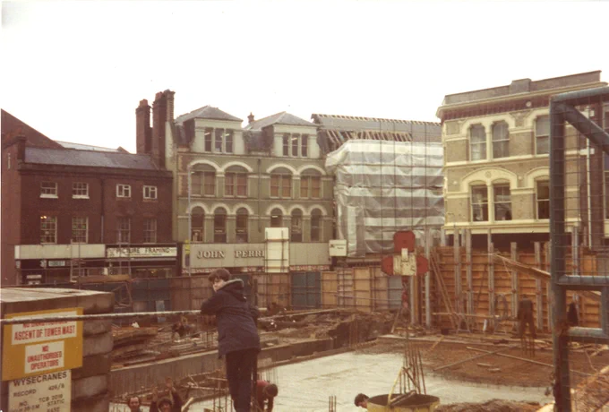 Construction site in front of buildings, with a person leaning on a barrier in the foreground.