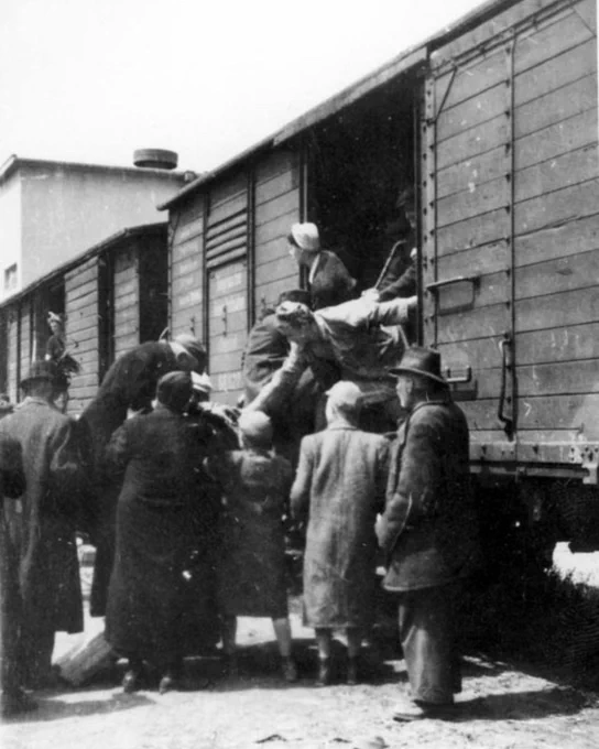 A group of people wearing early to mid-20th century clothing gather around a train car with open doorways. Several individuals are helping others to climb into the train car, which appears to be a freight type, with some onlookers observing the scene. Deportation of Jews from Hungary to Auschwitz.