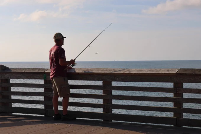 A person stands on fishing pier, fishing with a rod and reel, overlooking a calm sea under a partly cloudy sky.