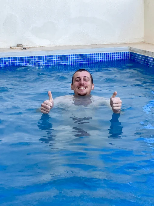 A smiling man gives two thumbs up while relaxing in a blue pool, surrounded by a light-colored wall.