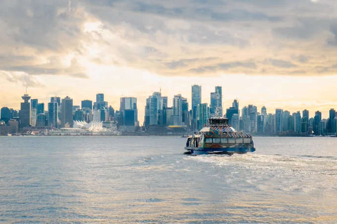 A SeaBus crossing calm waters with the Vancouver skyline under a cloudy sky in the background.