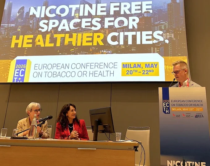 The image shows three individuals on a panel at the European Conference on Tobacco or Health in Milan from May 20th to 22nd, 2026. A large banner behind them reads "Nicotine Free Spaces for Healthier Cities." Two panelists are seated at a table with microphones, while Dr. Marc Bjurlin stands at a podium with conference branding.