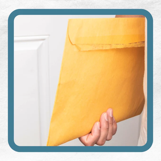 A hand holding a large yellow padded envelope in front of a white door.