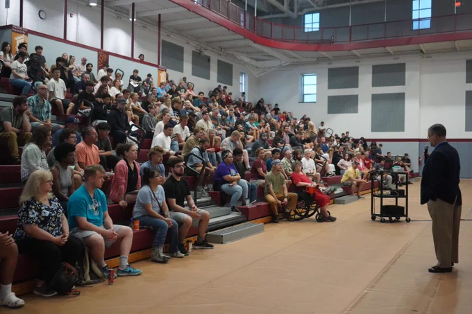 A large group of people seated on bleachers attentively listening to a speaker in a gymnasium setting.