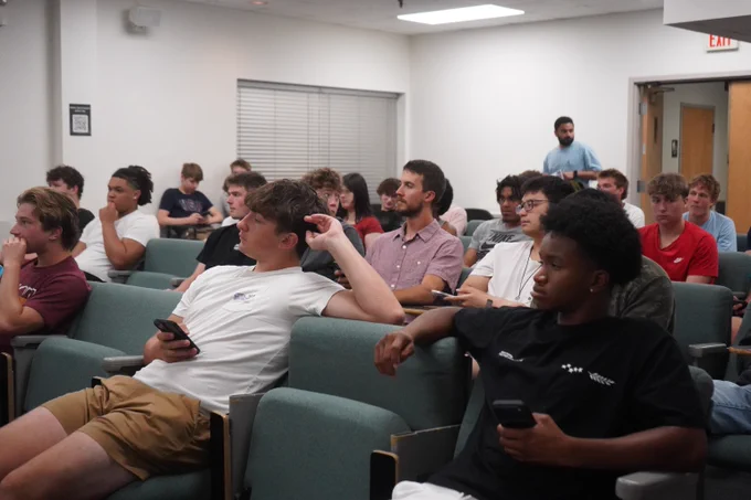 A classroom filled with students seated in rows, some holding phones.