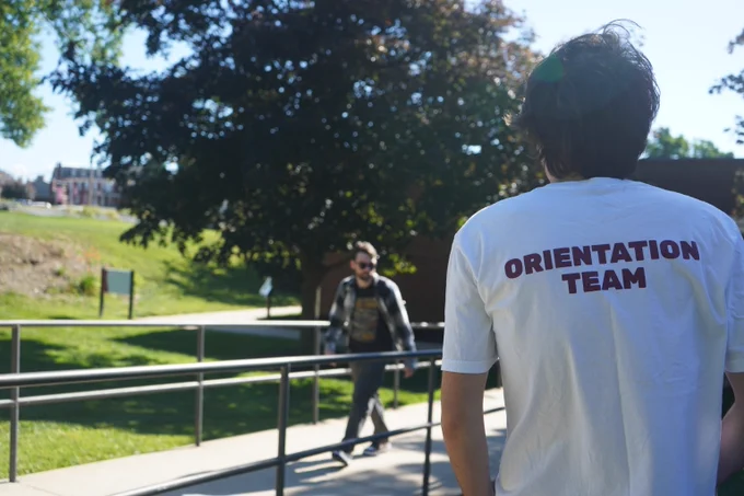 Person wearing a white shirt labeled 'Orientation Team' standing outdoors near a ramp with another person walking in the background on a sunny day.