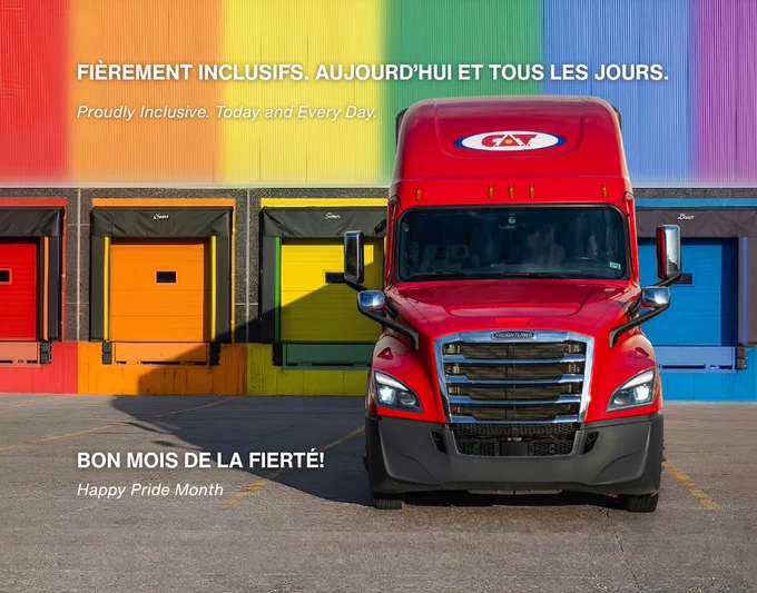 A bright red semi truck is parked facing forward in front of a warehouse with multiple loading dock doors painted in rainbow colors from red to purple. The building façade above the doors displays a gradient rainbow backdrop. White text on the image reads “Fièrement inclusifs. Aujourd’hui et tous les jours.” with the English translation “Proudly Inclusive. Today and Every Day.” In the lower left corner, additional white text reads “Bon mois de la fierté” and “Happy Pride Month.” The scene is well lit with sunlight casting shadows on the pavement.