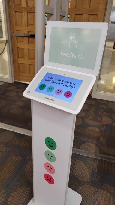 Podium sitting in front of the library’s exit with a touchscreen on top with four smiley faces from happy to angry, with the question prompt: “How happy are you with the HSCL today?”