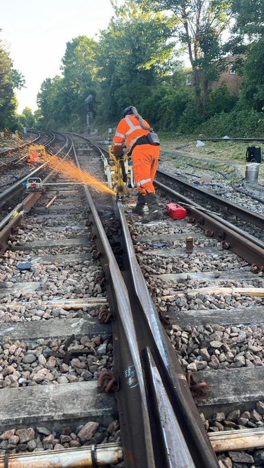 Rail worker in high-visibility orange PPE using a grinding tool on railway tracks, with bright sparks flying along the rail in a tree-lined area.