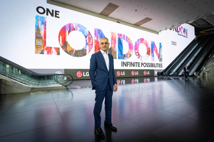 Mayor of London Sadiq Khan in a blue suit stands in a large, modern indoor exhibition/event space. Behind him is a massive white wall displaying the words “ONE LONDON” in large, colourful letters featuring images of London landmarks, along with the phrase “INFINITE POSSIBILITIES” underneath. 
