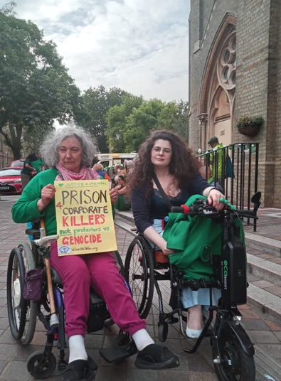 Two women wheelchair users side by side outside the Notting Hill Methodist Church building.  One holds a placard, Justice for Grenfell, Prison for corporate killers, not protesters against genocide.  winvisible.org The other holds a green coat (green for Grenfell).