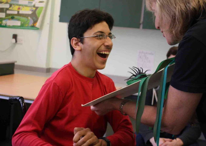 Teenager wearing a red shirt smiling at a woman holding out a book and telling a story. 
Image credit: Bag Books 