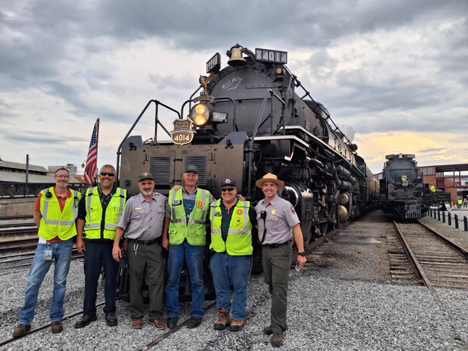 Six men, two park rangers and four train crew, stand in front of Big Boy 4014 at Steamtown. Big Boy 4012 sits to the back right of the photo.